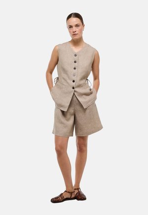 Femme portant un gilet beige sans manches boutonné et un short assorti avec des chaussures plates marron tissées, debout les mains dans les poches.