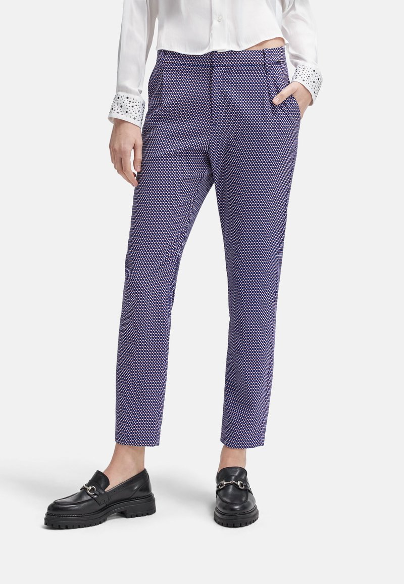 Femme portant un pantalon slim violet à motifs, des mocassins noirs, et un chemisier blanc avec des poignets à motifs, debout la main dans la poche.
