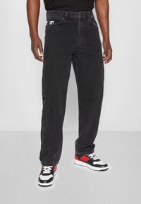 Man i lösa svarta jeans och svarta, vita och röda sneakers som står med avslappnade händer vid sidorna.