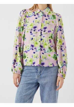 Kobieta ubrana w długą koszulę na guziki z zielono-fioletowym kwiatowym nadrukiem na różowym tle, zestawioną z jasnoniebieskimi dżinsami.