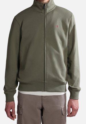 Uomo che indossa una giacca verde oliva con zip e un piccolo distintivo della bandiera norvegese sul petto, insieme a pantaloni cargo beige.