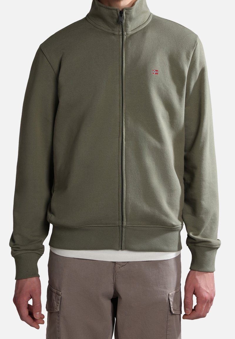 Uomo che indossa una giacca verde oliva con zip e un piccolo distintivo della bandiera norvegese sul petto, insieme a pantaloni cargo beige.