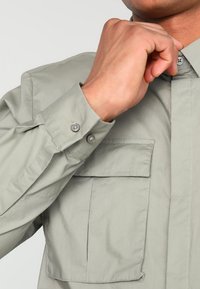 Osoba zapinająca górny guzik na jasnoszarej koszuli z długim rękawem, która ma kieszeń na piersi i zapięcie przy mankietach.