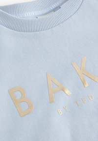 Világoskék pulóver puha anyagból, aranyfóliás "BAK BY TED BAKER" felirattal és bordázott nyakkal.