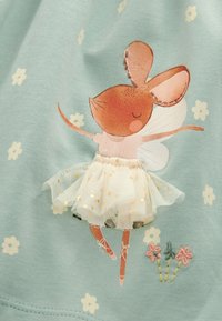 Souris brune dansante aux yeux fermés, portant un tutu blanc et des chaussures de ballet, sur un tissu bleu clair à petites fleurs blanches.