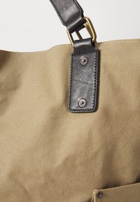 Canvas tote taske i khaki med en sort læderrem. Udstyret med messinghardware og en frontlomme med knaplukning. Struktureret overflade.