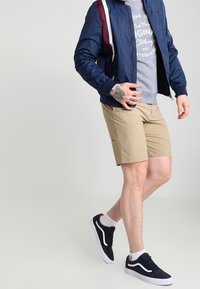 Uomo che indossa una giacca bomber blu navy, una maglietta grigia con stampa, pantaloni corti beige, calze bianche e sneakers nere, mostrando un tatuaggio sulla mano mentre cammina.