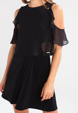 Woman wearing a black cold-shoulder dress with sheer flutter sleeves and a flared skirt against a neutral background.