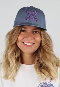 Casquette gris charbon avec broderie florale violette sur le devant ; visière courbée ; portée sur une chemise blanche avec un design graphique.