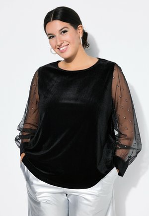 Vrouw lacht in een zwarte fluwelen top met doorschijnende polkadotmouwen en zilveren broek, handen in de zakken, tegen een witte achtergrond.
