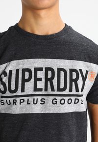 Šedé a tmavě šedé tričko s krátkým rukávem s kontrastním černým a bílým textem "SUPERDRY SURPLUS GOODS" v tučném písmu, s oranžovým logem jako akcentem.