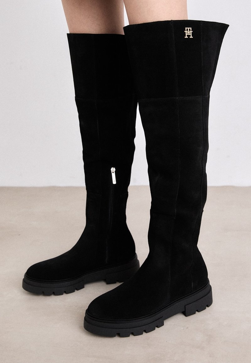 Bottes en suède noirs, au-dessus du genou, avec une semelle épaisse en caoutchouc noir. Elles disposent d'une fermeture éclair sur le côté et d'un accent logo doré en haut.