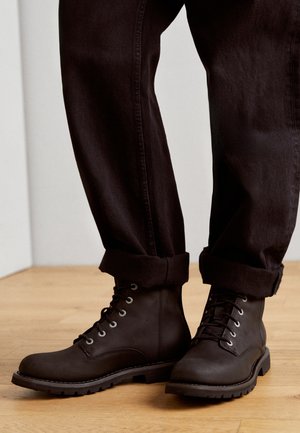 Botas marrones con cordones y ojales de metal, con punta redonda y superficie texturizada. Combinadas con pantalones oscuros de puños enrollados sobre un suelo de madera.