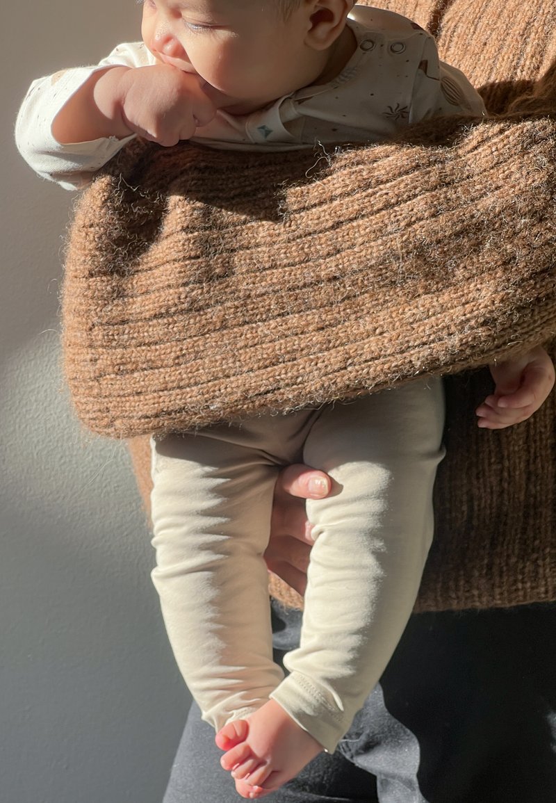 Pull en tricot marron avec une texture côtelée, tenant un enfant portant des leggings beige clair et pieds nus, dans une douce lumière naturelle.