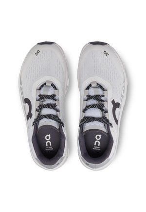 Valkoiset ja harmaat On Cloud Monster -juoksukengät mustavalkoisilla nauhoilla, kuvattuna ylhäältä vaalealla taustalla.