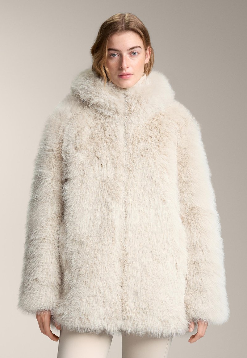 Kabát z umelého kožušiny svetložltej farby, s širokou kapucňou, voľného strihu a mäkkej, nadýchanej textúry, s rovnou lemovkou a spustenými ramenami.