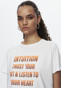 T-shirt en coton blanc avec un texte en dégradé orange audacieux : "INTUITION FAIS CONFIANCE À TON INSTINCT & ÉCOUTE TON COEUR." Manches courtes et col rond.