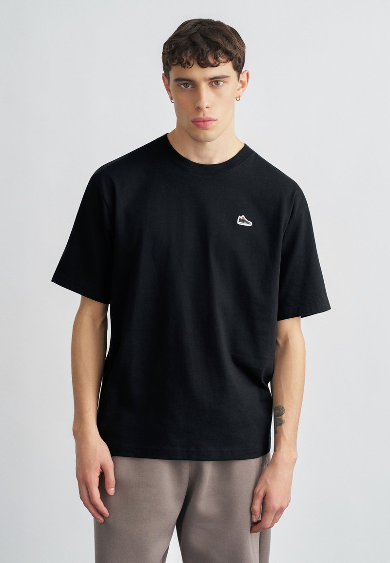 Μαύρο βαμβακερό T-shirt με χαλαρή εφαρμογή, που διαθέτει μικρό κεντημένο λογότυπο αθλητικού παπουτσιού στο στήθος και στρογγυλή λαιμόκοψη.