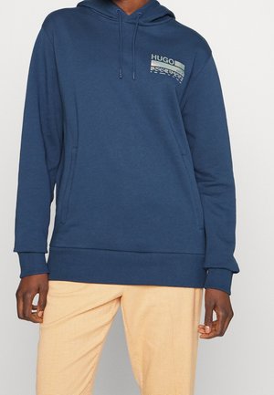 Osoba w niebieskiej bluzie z kapturem z logo „HUGO” na piersi i jasnobeżowych spodniach, stojąca na tle jednolitego tła.