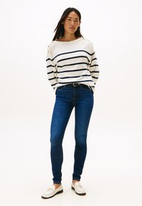 Wit gestreepte trui met blauwe strepen, schouderknopen, gecombineerd met donkerblauwe skinny jeans en witte loafers met gouden accenten.