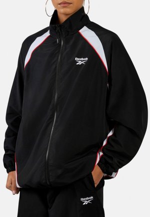Personne portant un survêtement noir Reebok avec des accents blancs et rouges, de grandes boucles d'oreilles en forme d'anneau et les mains dans les poches.