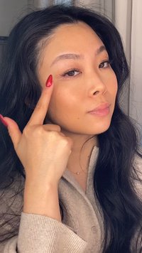 Młoda kobieta z długimi ciemnymi włosami wskazuje na swoją prawą brew pomalowanymi na czerwono paznokciami, ubrana w beżowy sweter i delikatny perłowy naszyjnik.