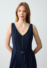 Abito blu navy con scollatura a V profonda, vita con cintura e gonna plissettata, completato da una delicata collana con pendente in oro e tessuto morbido.