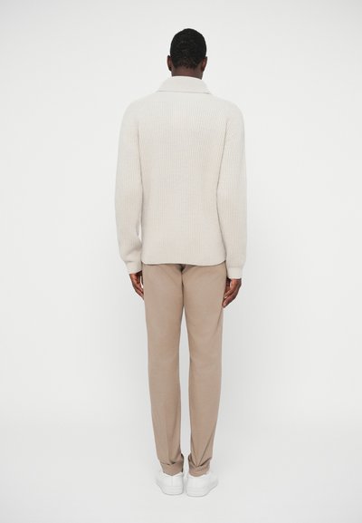 Beige kötött pulcsi széles gallérral, világosbarna nadrággal és fehér sportcipőkkel párosítva. Testhez álló ruházatban, hátulról látható.