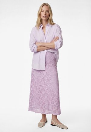 Femme debout, bras croisés, portant une chemise rayée violet clair, une jupe en dentelle lavande assortie et des ballerines beiges tissées à bride arrière sur fond blanc.