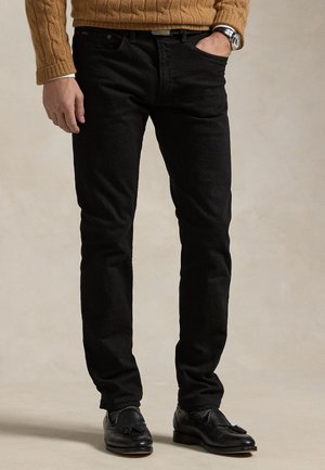 Homme portant un jean noir slim, des mocassins noirs à pampilles, un pull en maille torsadée marron et une montre-bracelet, une main dans la poche.