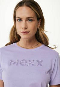 Fialové bavlněné tričko s flitrovaným logem "MEXX", kulatým výstřihem, krátkými rukávy a hladkou texturou. Zlatý řetízkový náhrdelník zdůrazňuje design.