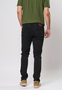 Jeans slim fit noirs avec une texture subtile, dotés de deux poches arrière et d'un patch en cuir marron. Associés à un polo vert et des baskets beiges.