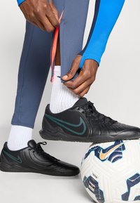 Nike svarte treningssko med grønne detaljer, grå treningsbukser med oransje glidelåser, og en hvit fotball med blå og oransje mønster.