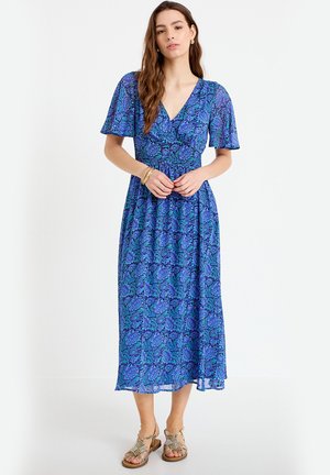 Vrouw in een blauw bloemenmidi-jurk met vleermuismouwen en V-hals, met gouden armbanden en gouden sandalen met bandjes, staand tegen een witte achtergrond.