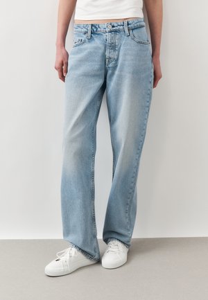 Person iført løstsiddende lyseblå jeans og hvide sneakers, der står mod en ensfarvet væg og gulv.