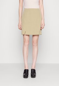 Jupe longueur genou, vert olive clair avec une texture lisse, présentant deux fentes latérales et un design structuré, assortie à des chaussures en cuir noir.
