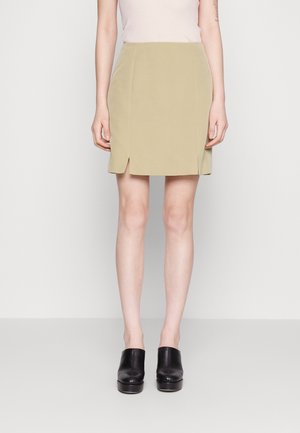 Jupe longueur genou, vert olive clair avec une texture lisse, présentant deux fentes latérales et un design structuré, assortie à des chaussures en cuir noir.