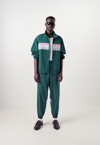 Fato de treino verde feito de material leve com riscas verticais em rosa, roxo e azul. Usado com sapatos pretos e óculos de sol.