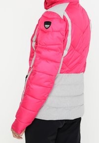 Puffer jacket featuring bright pink upper and gray lower sections, quilted texture, and a logo patch on the shoulder. Zippered front pockets.