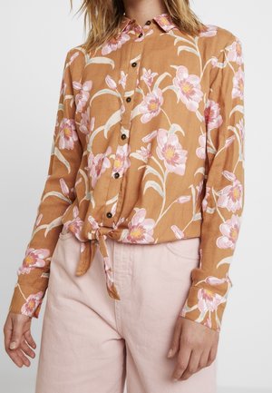 Femme portant une chemise marron à boutons avec imprimé floral rose nouée à la taille, associée à un pantalon rose clair taille haute.