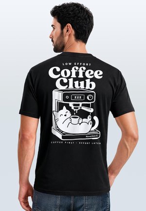 Mann trägt schwarzes T-Shirt mit weißer Grafik einer Katze, die entspannt in der Kaffeemaschine sitzt und eine Tasse hält, Text "Low Effort Coffee Club" und "Coffee first effort later."