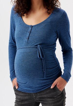 Personne enceinte portant un haut bleu à manches longues avec des boutons et une ceinture latérale, mains dans les poches d'un jean gris foncé.