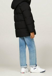 Osoba v černé zateplené bundě, světlých modrých kalhotách se širokými nohavicemi a bílých teniskách s modrými a červenými detaily, stojící na neutrálním pozadí.