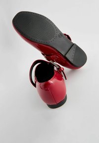Rote Lacklederschuhe mit niedrigem Absatz, Knöchelriemen und strukturierten schwarzen Gummisohlen. Sichtbares Schnallen-Detail und glatte Oberfläche.