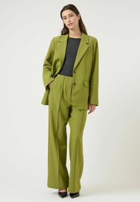Blazer vert et pantalon à jambes larges assorti en tissu texturé. La tenue comprend un haut gris et des chaussures noires pointues.
