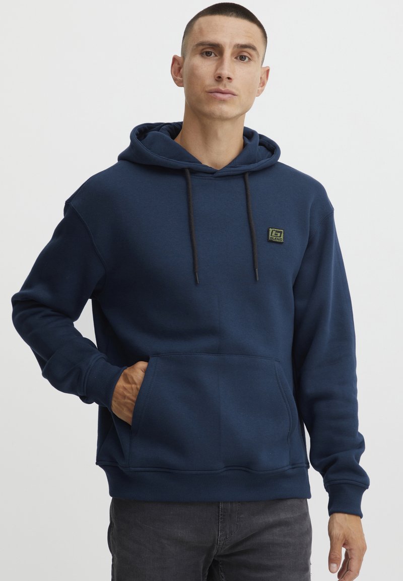 Marinblå hoodie med en framficka, justerbar snörning på huvan och en liten grön logotyp på bröstet. Tillverkad av mjukt tyg med ribbade ärmslut.