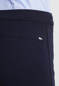 Marinblå byxor i mjukt material med ett diskret bakfick och en liten silverlogotyp. Slät yta med minimal sömnad.