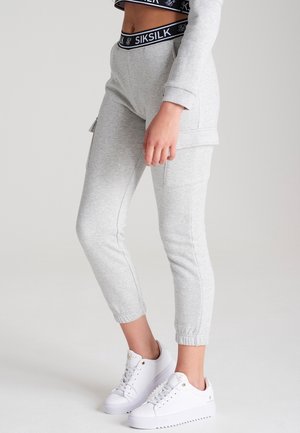 Nærbillede af en person iført grå joggingbukser med cargolommer, en sort linning med mærket "SIKSILK" og hvide platåsneakers.