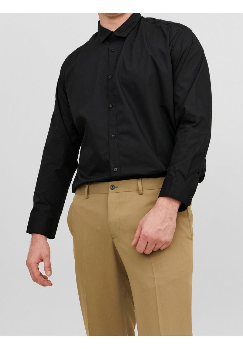 Homme portant une chemise noire à manches longues boutonnée, rentrée dans un pantalon beige, debout devant un fond clair uni.