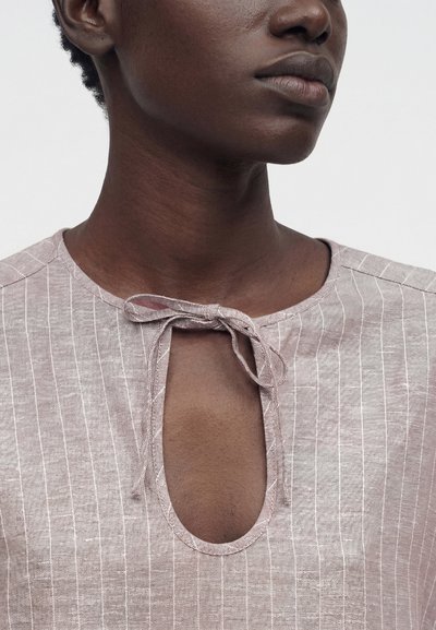 Κοντινό πλάνο ενός ατόμου που φοράει ριγέ τοπ ανοιχτού μωβ χρώματος με λαιμόκοψη σε σχήμα κλειδαρότρυπας, δεμένη με φιόγκο στον γιακά.